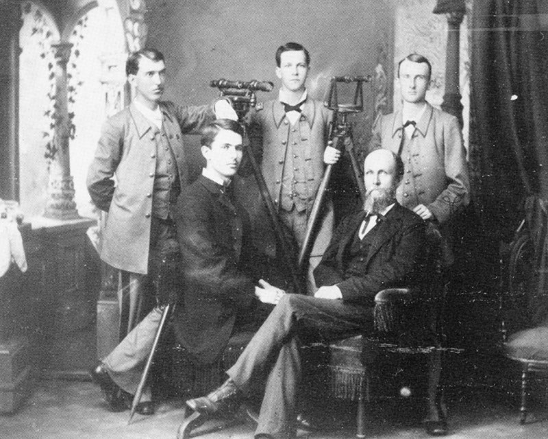Old picture of five men, two seated.